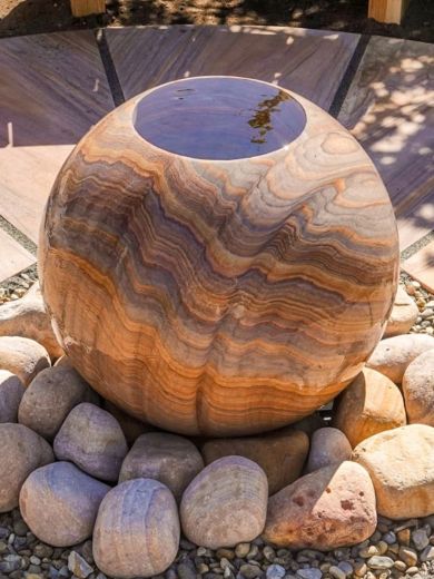 A sphere rainbow sandstone water feature with an infity top, surrounded by tumbled sandstone pebbles. 